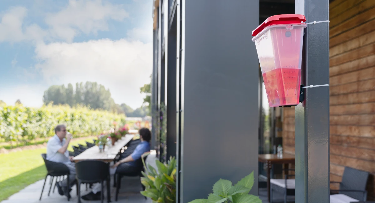 A close-up of a red and clear EcoCatch fly trap mounted to a pillar on an outdoor restaurant patio, with customers dining at a long table in the blurred background