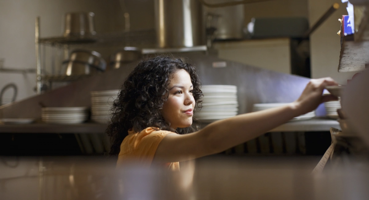 A waitress takes a plate from a gantry in a kitchen environment