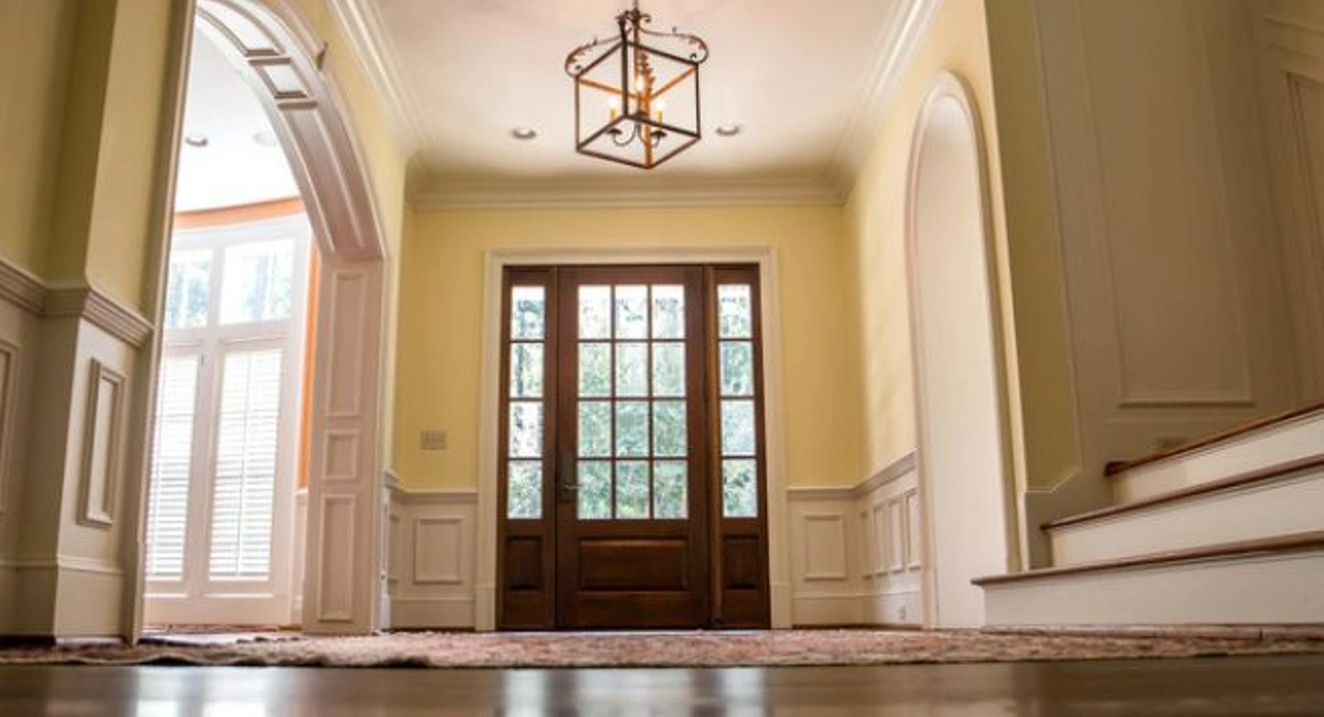 Grand and elegant yellow entrance to a home with wood floors and a staircase to the right of the wooden front door. 